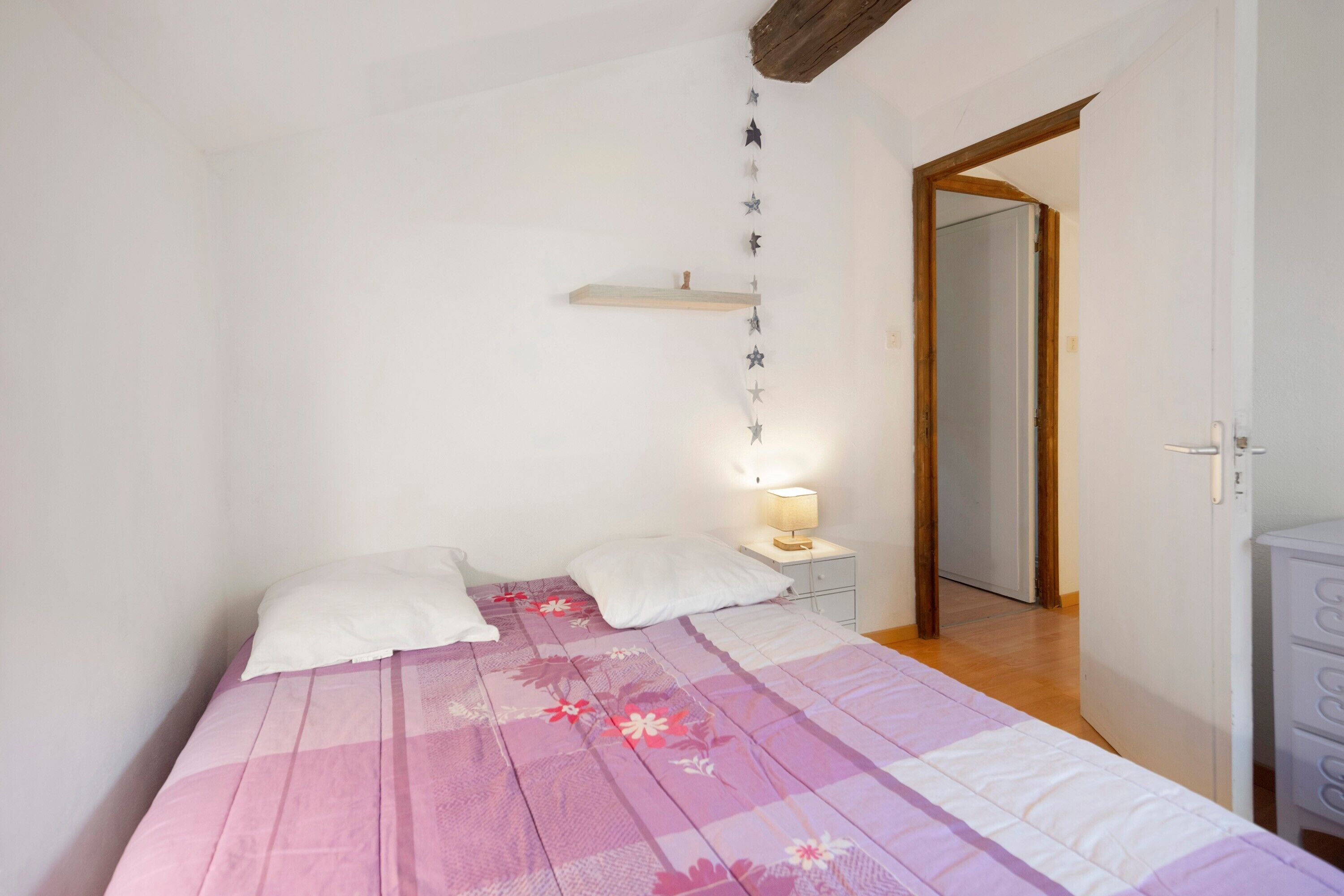 Une chambre à coucher avec un lit, une table de chevet avec une lampe, une poutre en bois au plafond et une porte donnant sur une autre pièce.