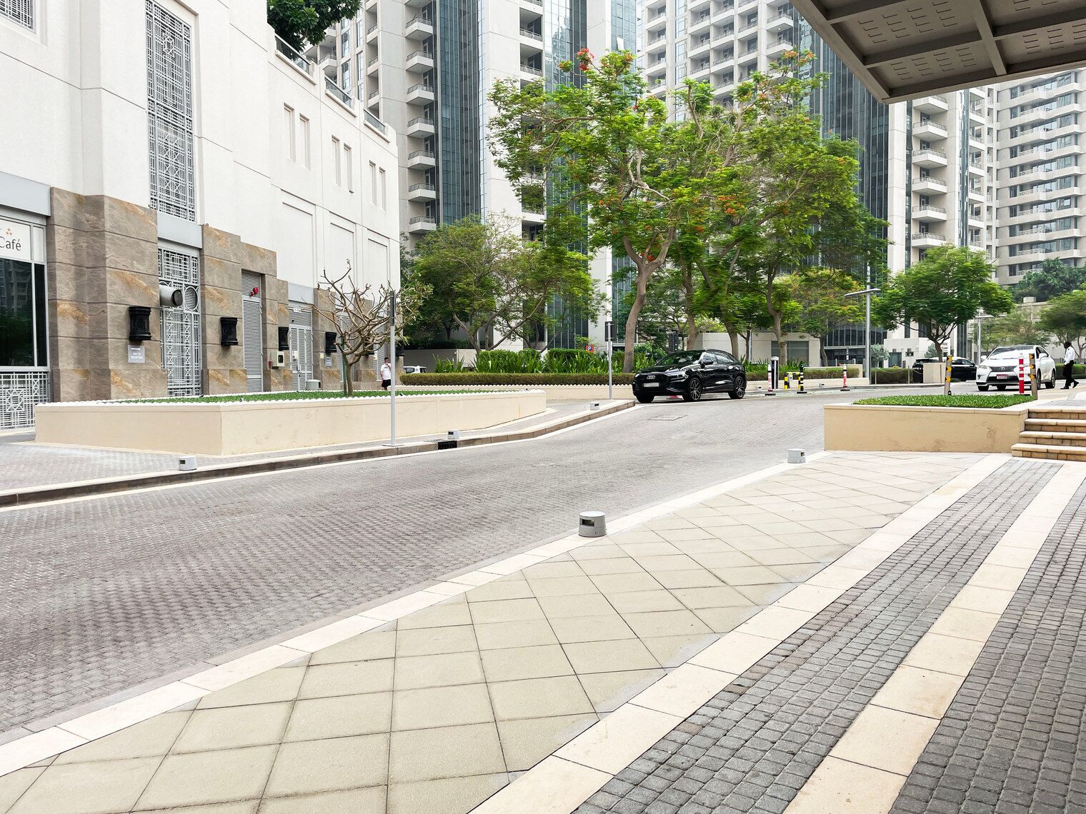 A modern urban street with high-rise buildings, a café, and a tree-lined sidewalk.