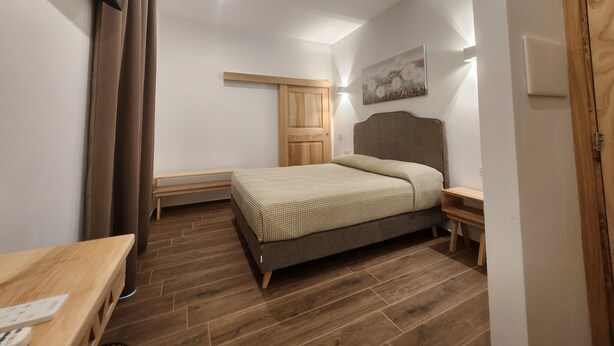 Une chambre à coucher comprenant un lit, une tête de lit en bois, des tables de chevet, un plancher en bois, un téléviseur fixé au mur et une porte.