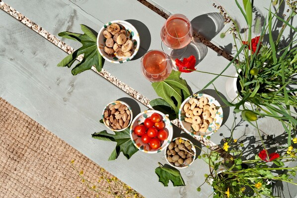 A table with bowls of nuts, cherries, and dried fruits, and glasses of a pink beverage, set outdoors with plants and flowers around.