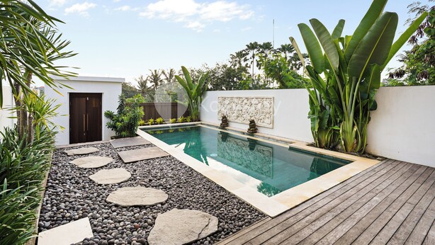 Una piscina rectangular con una terraza de madera y piedras que conducen a una puerta.