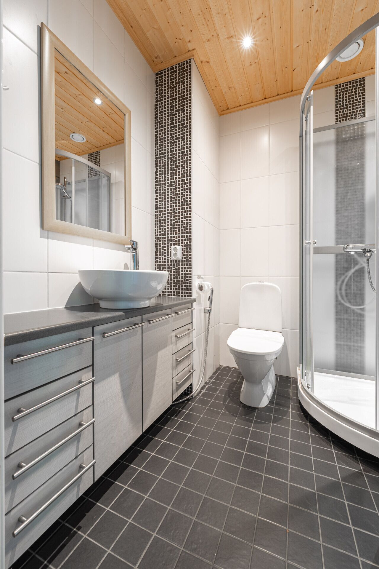Ein modernes Badezimmer mit Holzboden, einer in Glas eingefassten Dusche, einer weißen Toilette und einem großen Spiegel über dem Waschbecken.