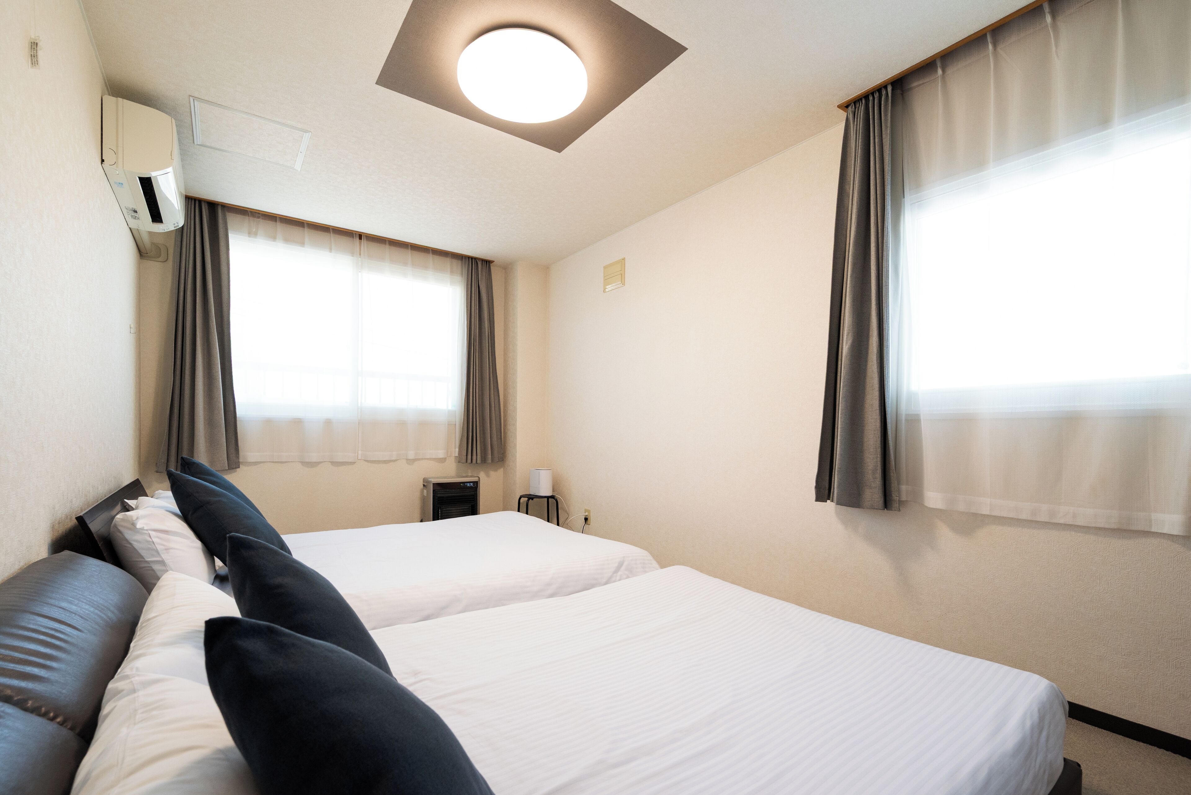 Ein Hotelzimmer mit zwei Betten, einer Deckenleuchte, einer Klimaanlage und Vorhängen an den Fenstern.
