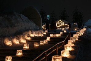 Área externa nevada com degraus que levam a um edifício, iluminada por velas.