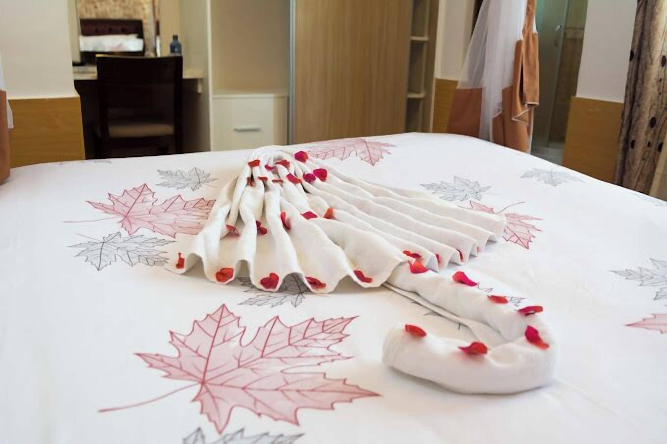 A neatly made bed with a white bedspread featuring leaf patterns and a towel arranged in a fan shape.
