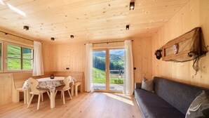 Ein Holzblockhaus-Innenraum mit Esstisch, Stühlen, einer Couch und einem großen Fenster mit Blick ins Freie.