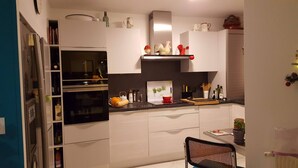 Cozinha compacta com armários brancos, forno preto e um pequeno espaço de refeições com uma cadeira.