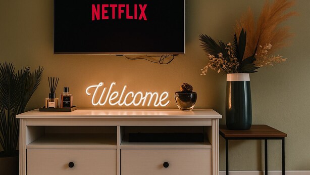 Seinälle asennettu televisio, jossa näkyy Netflixin logo, neonvaloin valaistu «Tervetuloa»-kyltti, kuivista kasveista tehty asetelma maljakossa sekä pieni pöytä, jonka päällä on kynttilä.