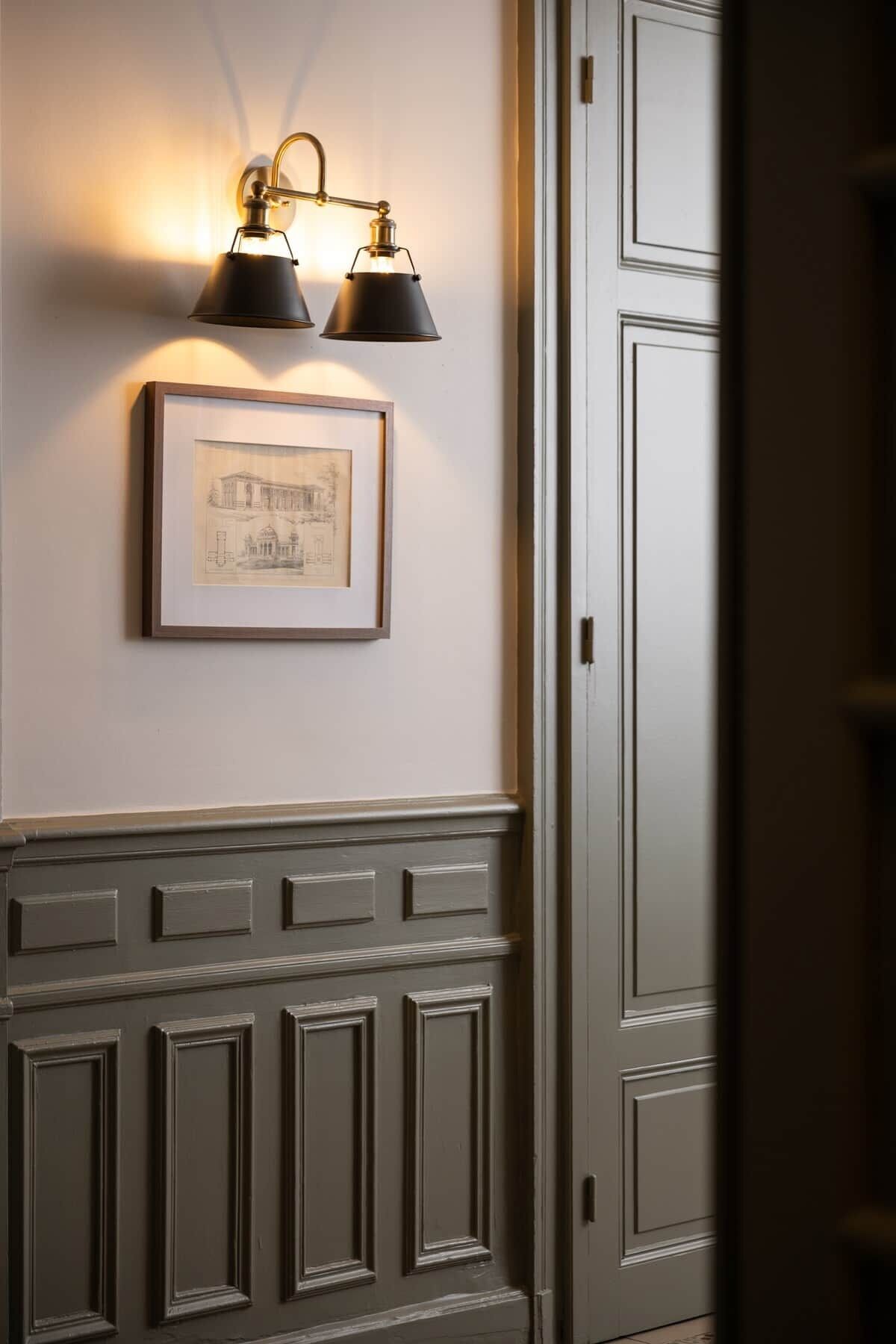 Ein Flur mit einem gerahmten architektonischen Entwurf an der Wand, einer Tür mit Griff und einer Wandleuchte mit zwei Lampen.