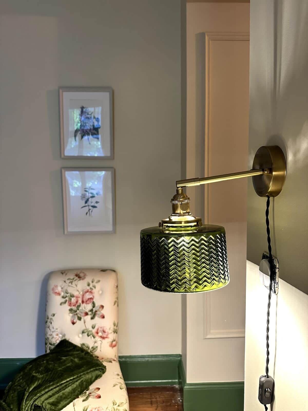 Een kamer met een groene lamp met structuur aan een muur, een fauteuil met bloemenprint en twee ingelijste foto's erboven.