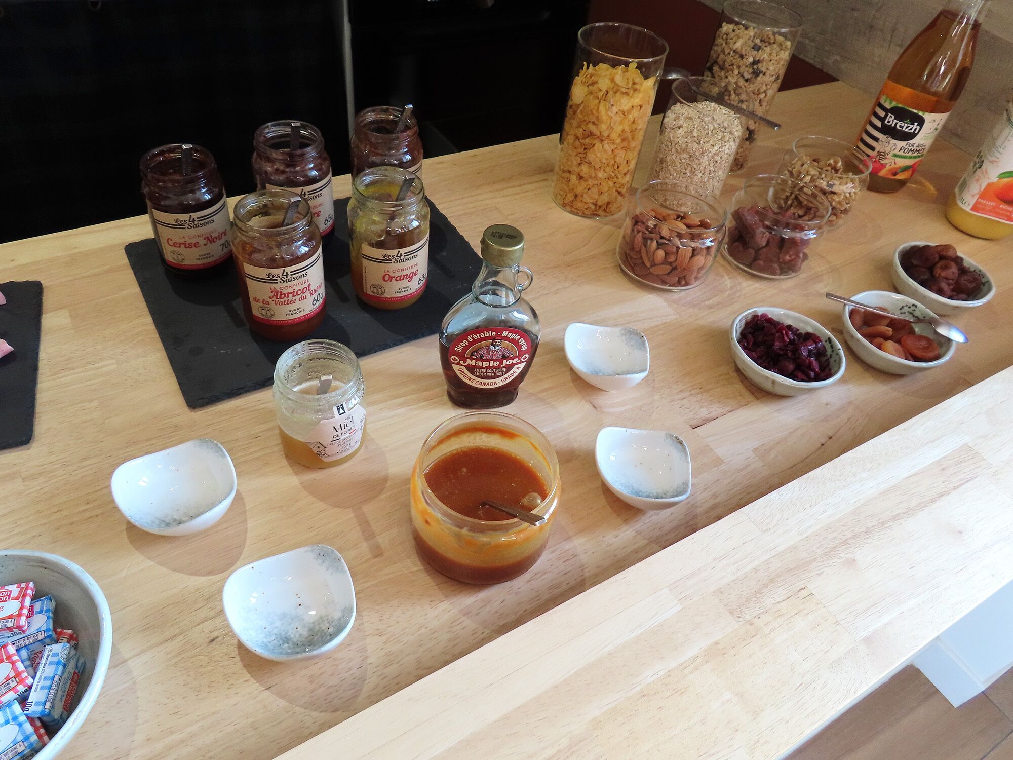A wooden table with various food items and containers, including jars of preserves, bottles of syrup, and bowls of nuts and dried fruits.