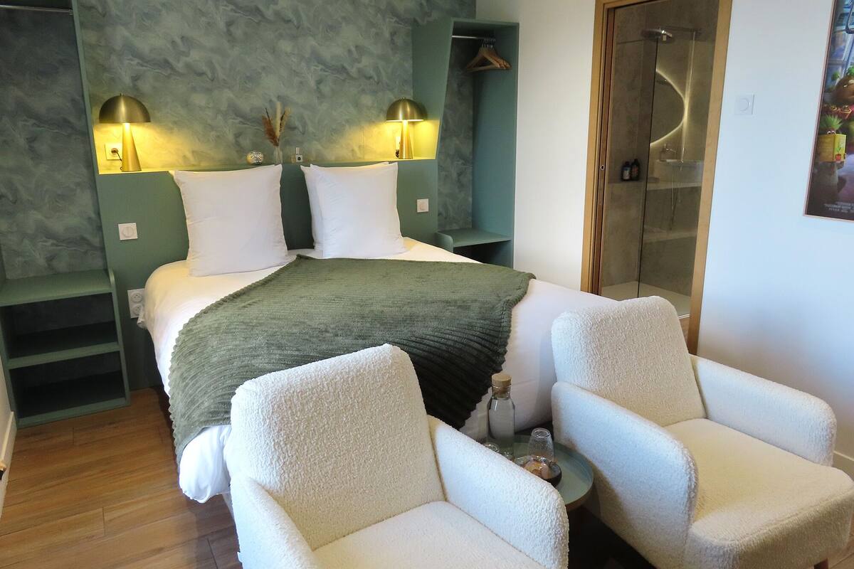 A bedroom with a bed, two bedside lamps, a green wall, a wooden door, and two white armchairs.