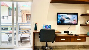 A room with a TV displaying 'HEROPANTIP 2', a desk with a computer, a chair, and a small table with a vase. - Stay near - Hotel near me Gurgaon (Gurugram)