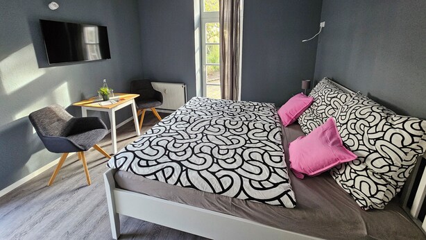 Quarto moderno com cama, duas cadeiras, uma mesinha e uma televisão.