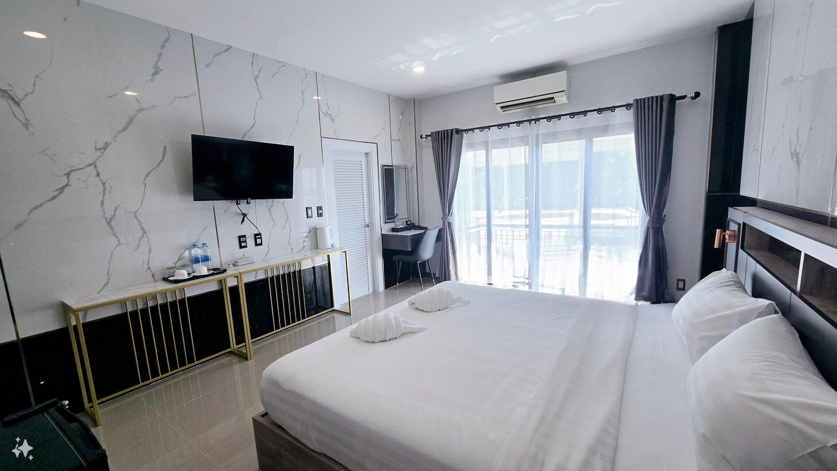Habitació d'hotel amb un llit gran, un televisor penjat a la paret, una taula de nit amb cadira i una finestra amb cortines.