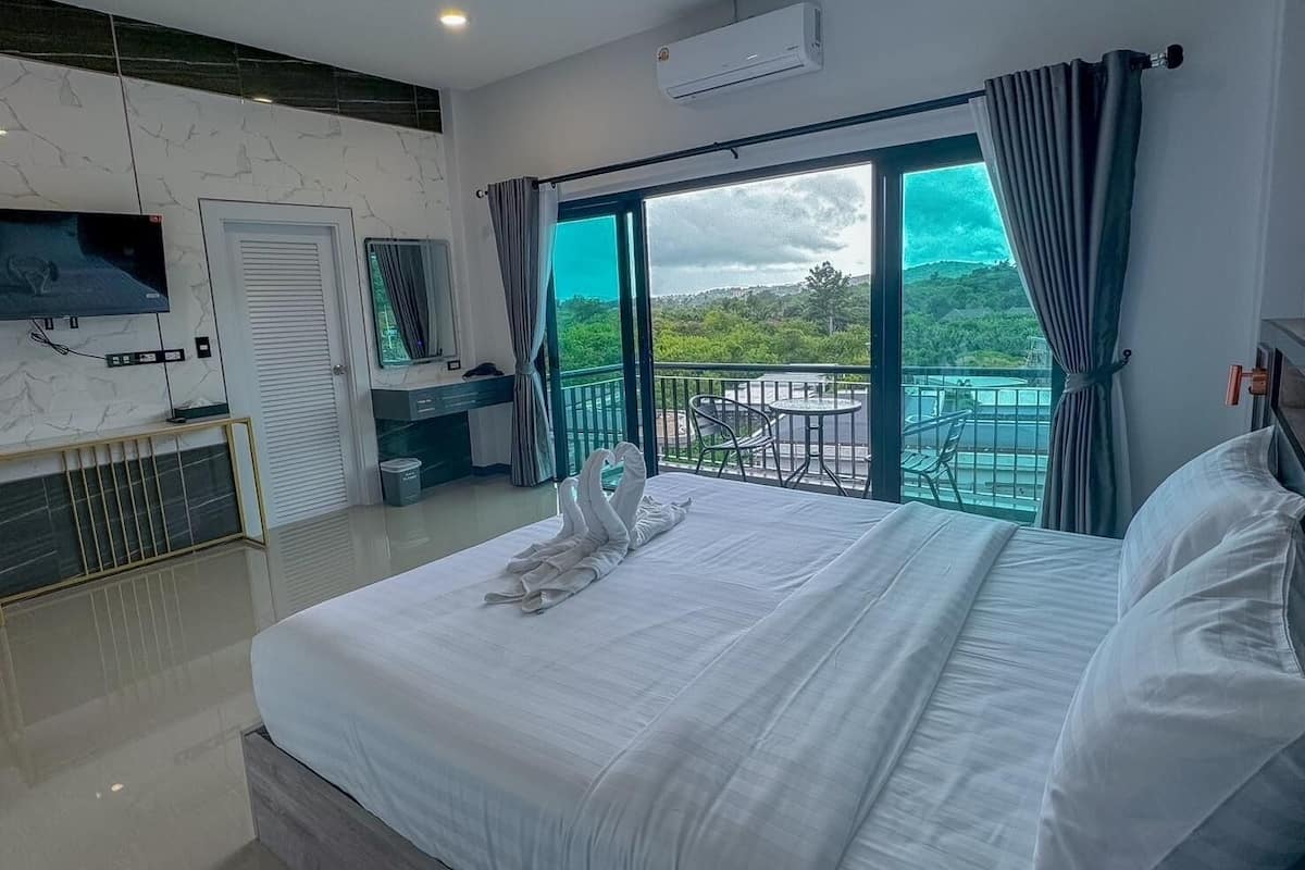 Une chambre d’hôtel moderne dotée d’un grand lit, d’un téléviseur fixé au mur, d’un balcon offrant une vue sur la verdure, ainsi que d’une petite table et d’une chaise.