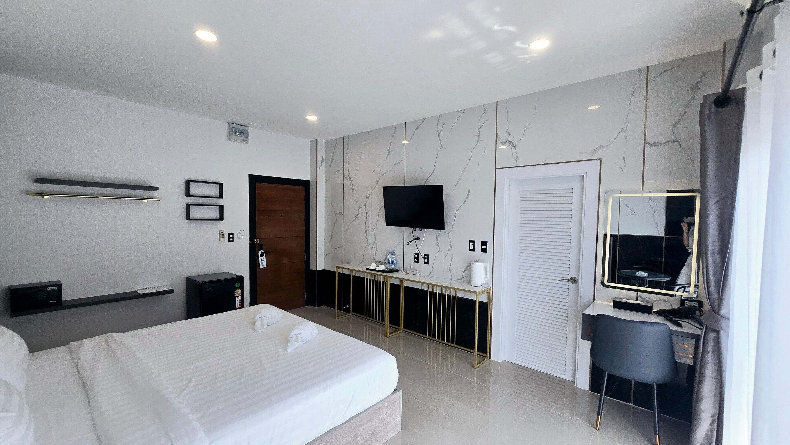 A modern hotel room with a large bed, a desk with a computer, a TV, and a bathroom with a mirror.