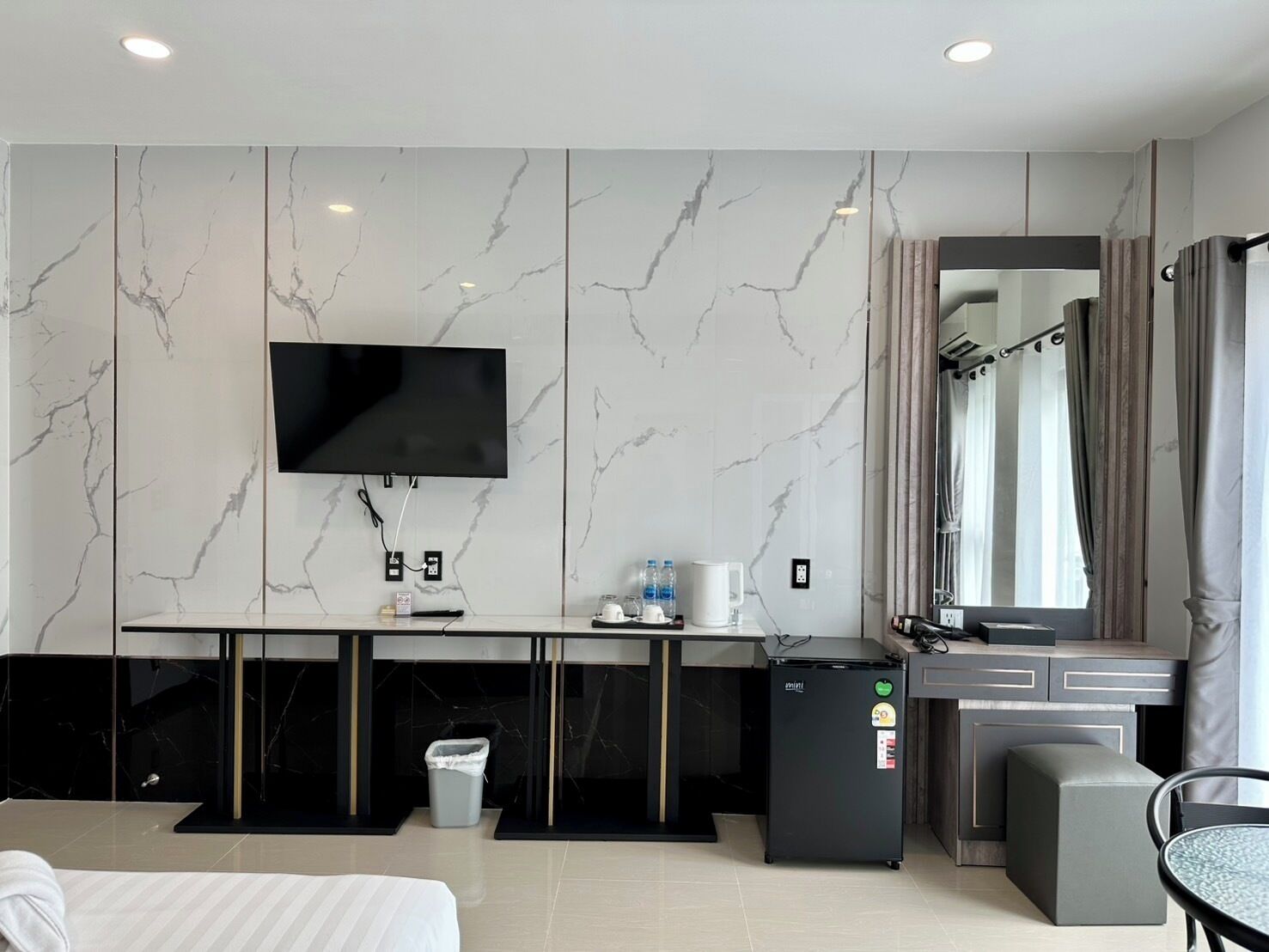A modern hotel room with a large bed, a desk with a computer, a small fridge, a TV mounted on the wall, and a bathroom with a mirror and curtains.