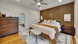 A bedroom with a bed, wooden panel wall, ceiling fan, bedside tables, and a dresser.
