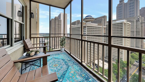 A balcony with a view of high-rise buildings, a potted plant, and a blue patterned rug.
