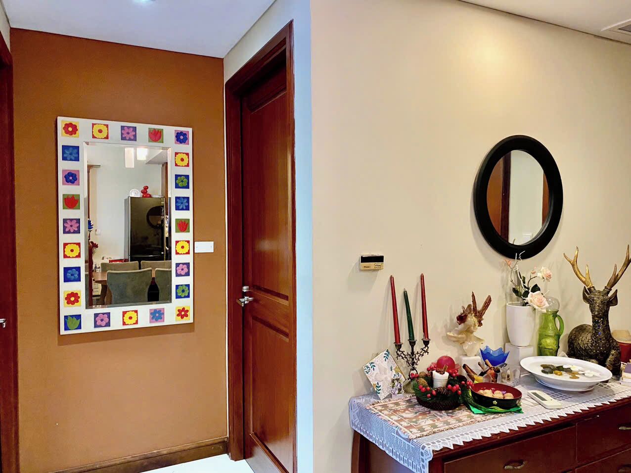 A room with a wooden door, a mirror with a colorful frame, and a table with various decorative items.