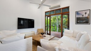 Ein modernes Wohnzimmer mit weißen Möbeln, einem Flachbildfernseher, einem Deckenventilator und einem großen Fenster mit Blick auf Grünflächen.