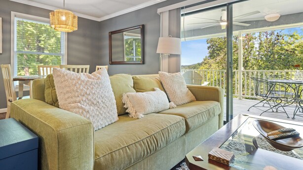 Ein grünes Sofa mit weißen Kissen, ein Glastisch und Blick auf eine Terrasse mit Gartenmöbeln.