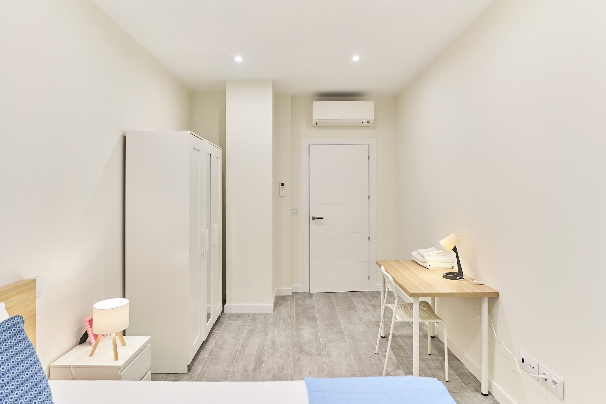 Une chambre d’hôtel de taille réduite, comprenant un lit, une table de chevet, une lampe, une armoire, un bureau, une chaise et un appareil de climatisation.