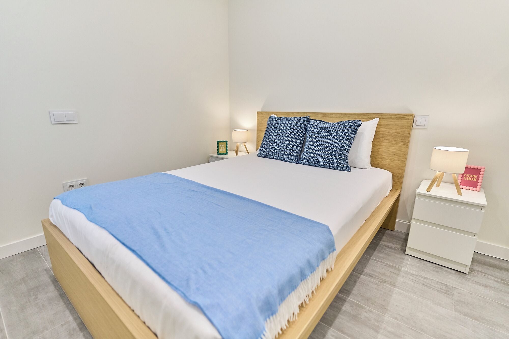 Una cama bien hecha con una colcha azul, un cabecero de madera y dos almohadas estampadas. Hay una mesita de noche con una lámpara y un pequeño objeto decorativo. La habitación tiene paredes blancas y un suelo de tono claro.