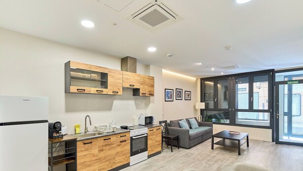 A modern kitchen with wooden cabinets, a built-in oven, and a microwave. There is a dining area with a table set for two, a sofa, and a window with a view of the street. - Apartamento Catedral Palace by gaiarooms (Valladolid)