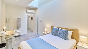 A modern hotel room with a large bed, a desk, and a chair. There is a wardrobe with a mirror and a hanging rack. The room has a skylight and a view of the hallway. - Apartamento Catedral Palace by gaiarooms (Valladolid)