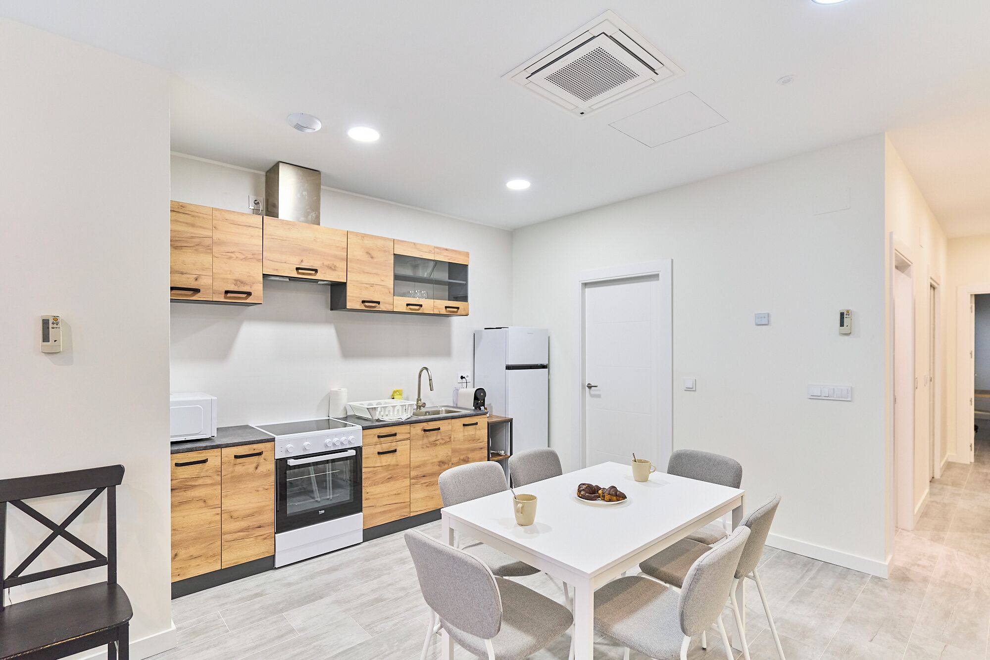 A modern kitchen with wooden cabinets, a white countertop, and a built-in oven. There is a dining area with a white table and grey chairs.