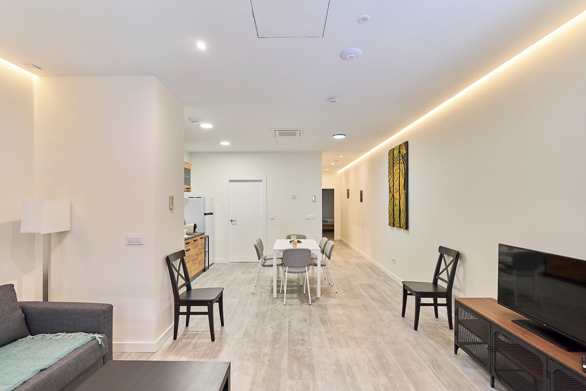 A modern living room with a sofa, a dining table with chairs, a TV, and a kitchen area in the background.
