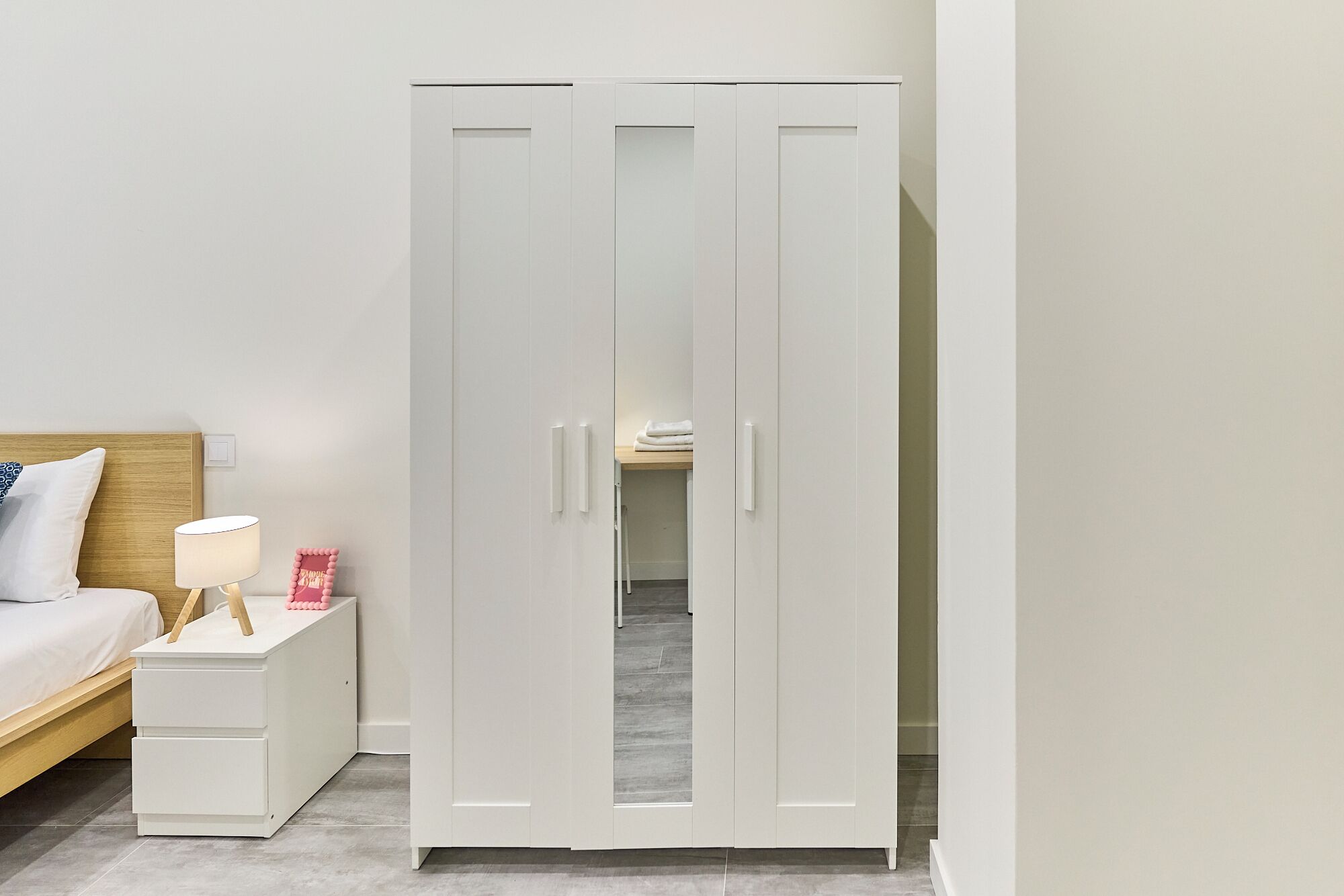 Um guarda-roupa branco com duas portas e uma pequena mesa de cabeceira com uma lâmpada e objetos decorativos.
