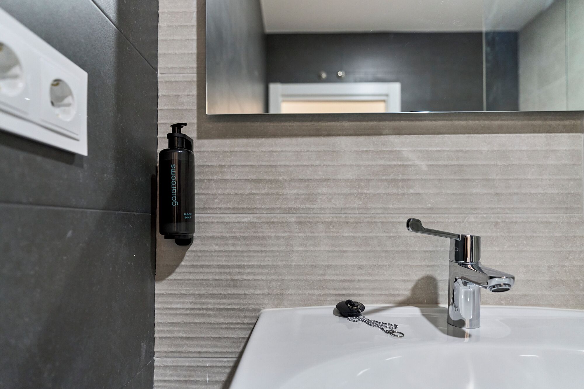 A bathroom with a white sink, a chrome faucet, and a soap dispenser on the wall.