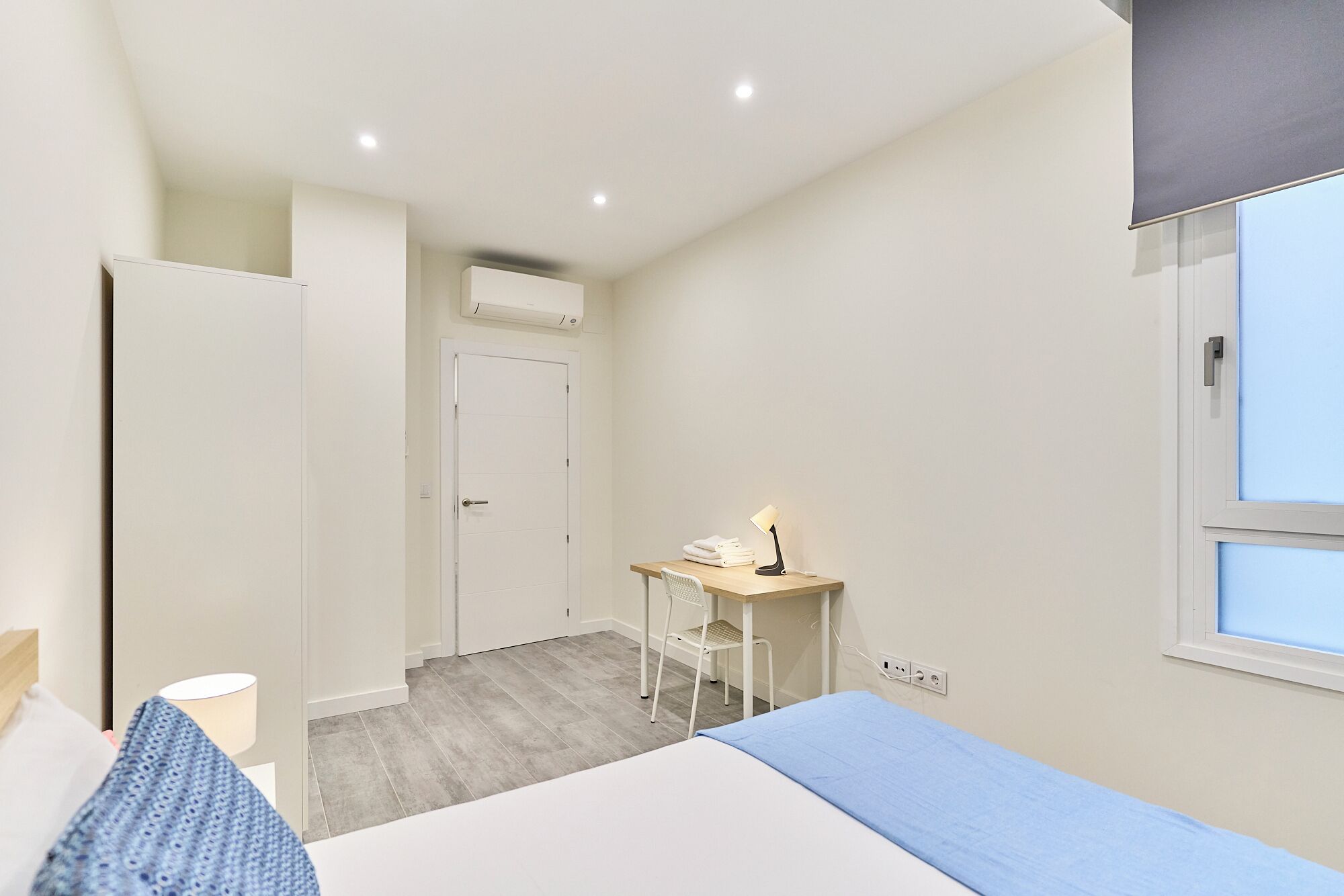 A bedroom with a bed, a desk with a chair, a window, and an air conditioning unit.