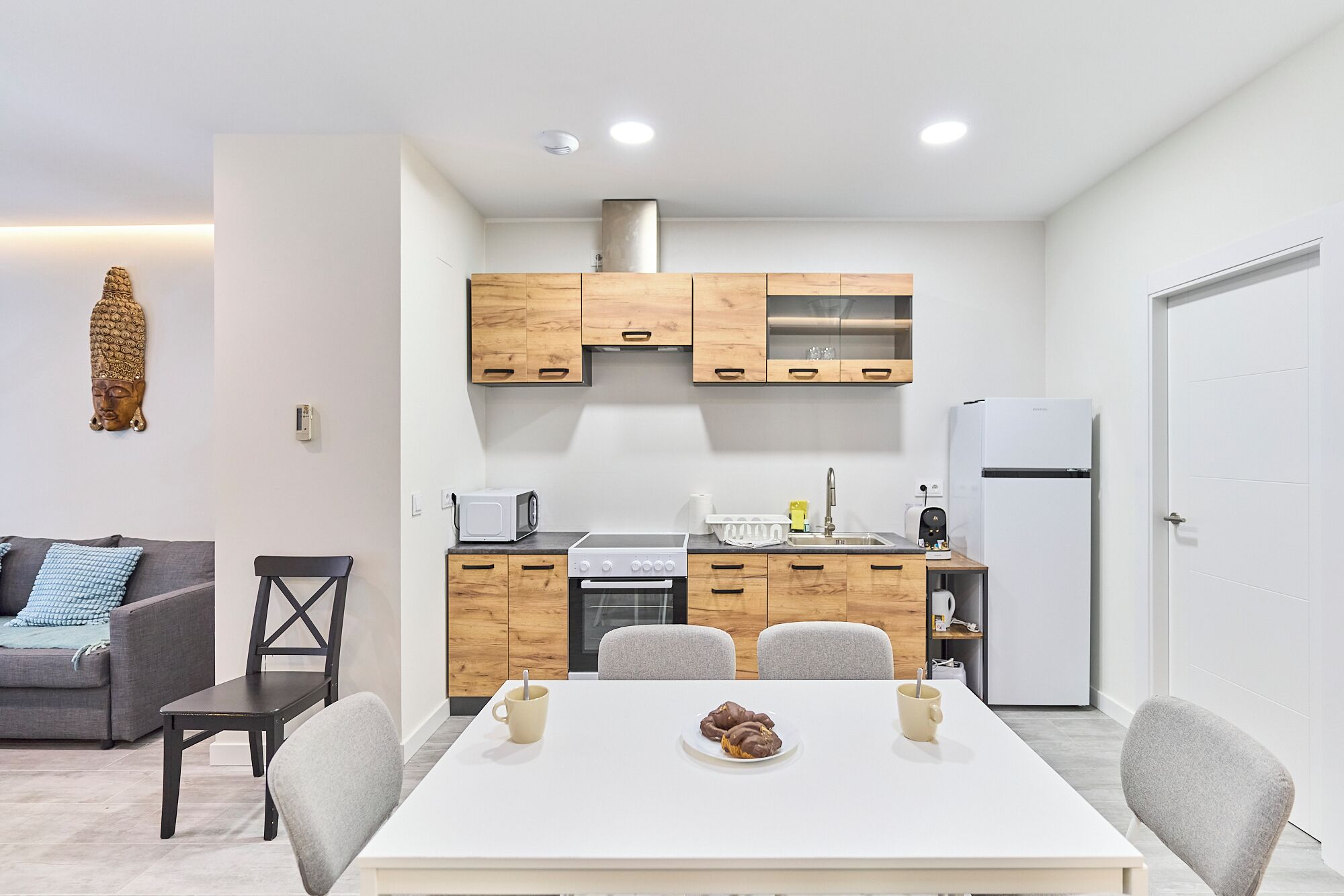 A compact kitchen with wooden cabinets, a microwave, a stove, and a refrigerator. There is a dining table with chairs and a small decorative Buddha statue on the wall.