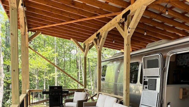 A covered outdoor deck with wooden flooring, a grill, wicker furniture, and a camper parked inside. - “Moonbeam” (Airstream) (Oakland)