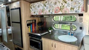 A compact kitchen with a microwave, stove, and sink, wooden cabinets, and a floral backsplash. - “Moonbeam” (Airstream) (Oakland)