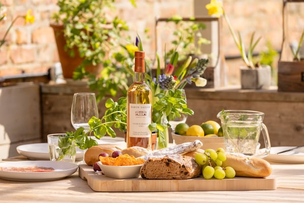 Une table dressée avec une bouteille de vin, du pain, des raisins et une carafe d’eau, entourée de plantes en pot et d’un mur de pierres rustiques.