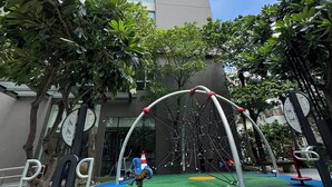 A playground with a swing set, climbing structures, and a slide, surrounded by trees and a building in the background.