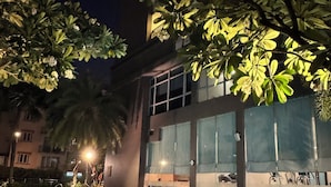 A nighttime pool area with a wooden deck, a swimming pool, and a building with large windows and a covered outdoor seating area.