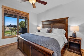 A bedroom with a large bed, wooden headboard, two bedside lamps, and a view of a green landscape through large windows. - Brasada Ranch Cabin 21 Mtn & Golf Views Hot Tub (Powell Butte)