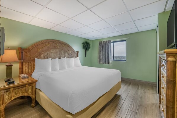 A hotel room with a large bed, a wooden nightstand with a lamp, a television, and a window with a view.