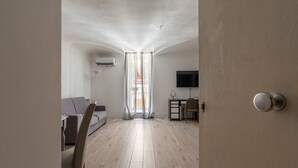 A modern living room with a wooden floor, a grey sofa, a dining table with chairs, a TV on a stand, and a window with sheer curtains. - Real Umberto I - Suite 1 by Wonderful Italy (Palermo)