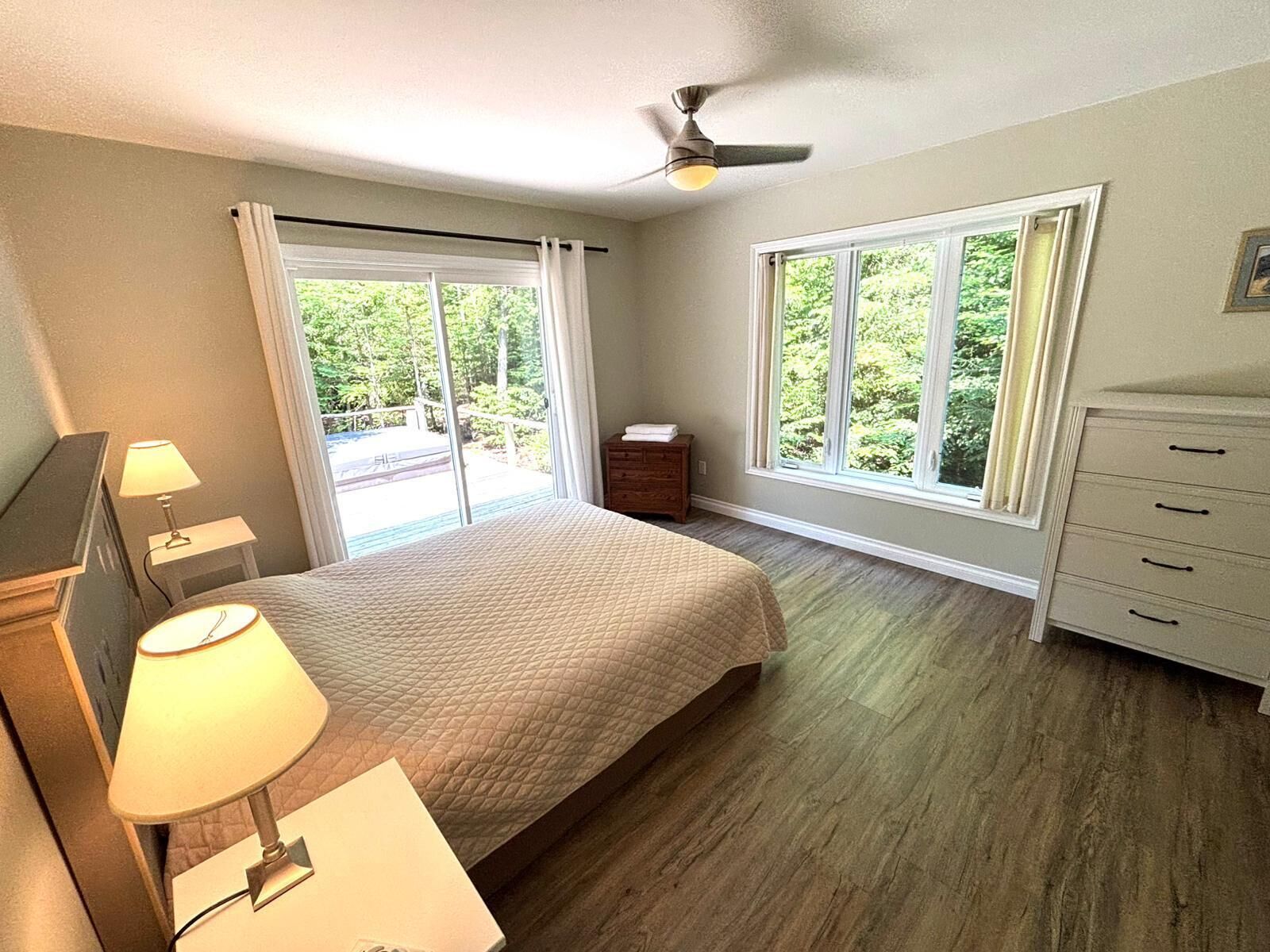 Quarto com cama, mesa de cabeceira, cômoda e ventilador de teto. Duas janelas oferecem vista para o exterior.
