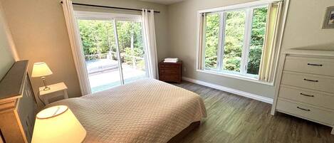 Quarto com cama, mesa de cabeceira, cômoda e ventilador de teto. Duas janelas oferecem vista para o exterior.
