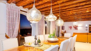 Un espace repas avec une table en bois, des chaises blanches, des lampes suspendues et une vue sur l’extérieur.