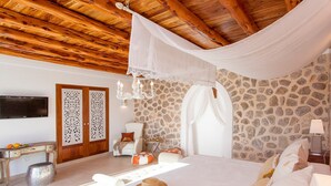Une chambre avec un mur en pierre, un lit avec une literie blanche, une télévision, un panneau décoratif au mur et un lustre.