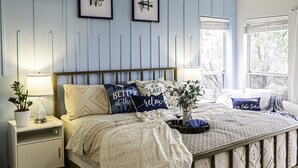 Une chambre avec un plafond en pente, un lit avec une tête de lit, deux tableaux encadrés au mur, une table de chevet avec une lampe et une fenêtre avec des rideaux.